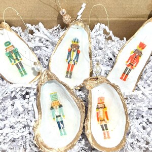 Op de afbeelding: Vijf met goud geverfde oesterschelpen met een wit interieur, elk met een ander notenkrakerontwerp. De notenkrakers zijn in verschillende kleuren geschilderd, waaronder rood, groen, blauw en goud.