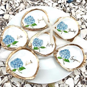 May include: Six white oyster shell dishes with gold rims, each featuring a hand-painted blue hydrangea and a name in cursive. A white starfish rests between the shells. The shells are arranged on a white plate, surrounded by shell fragments.