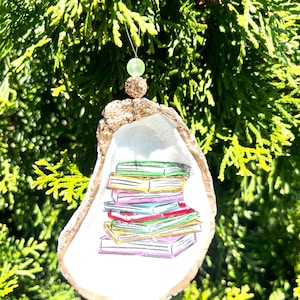 May include: A white oyster shell ornament with a gold rim, decorated with a stack of colorful books. The ornament is hanging from a green bead.