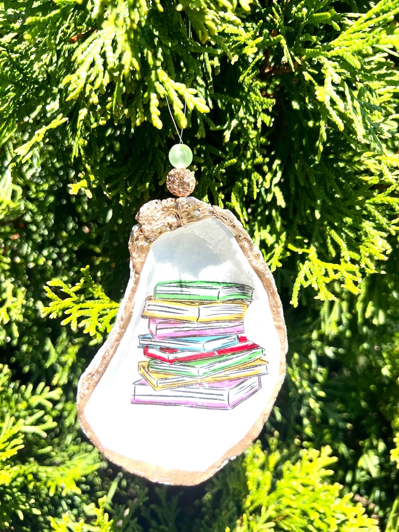 May include: A white oyster shell ornament with a gold rim, decorated with a stack of colorful books. The ornament is hanging from a green bead.