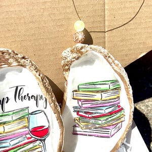 May include: Two oyster shells painted with gold trim. One shell features a stack of colorful books with the text "Book Therapy". The other shell features a glass of red wine and a stack of colorful books.