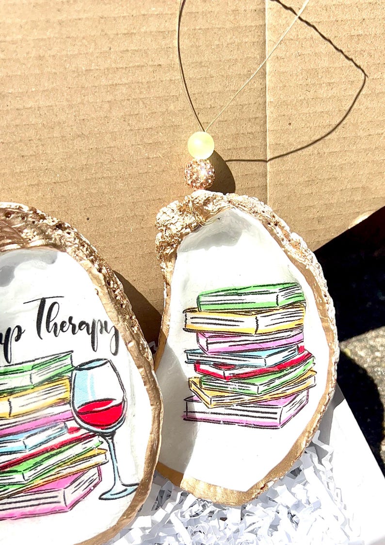 May include: Two oyster shells painted with gold trim. One shell features a stack of colorful books with the text "Book Therapy". The other shell features a glass of red wine and a stack of colorful books.