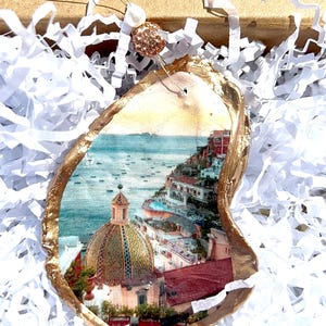 May include: An oyster shell ornament with a gold-painted edge. The shell features a scenic image of a coastal town with a domed building, red-roofed buildings, and a blue sea. The ornament is displayed on white paper shreds.