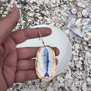 K&ouml;nnte beinhalten: Ein Anh&auml;nger bestehend aus einer wei&szlig; bemalten Austernschale mit einem blauen Fischmotiv und goldenem Rand, befestigt an einer d&uuml;nnen goldfarbenen Metallkette. Das Fischdesign zeigt komplexe florale Muster in Blau und Gelb auf dem K&ouml;rper.