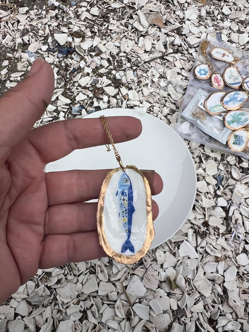 K&ouml;nnte beinhalten: Ein Anh&auml;nger bestehend aus einer wei&szlig; bemalten Austernschale mit einem blauen Fischmotiv und goldenem Rand, befestigt an einer d&uuml;nnen goldfarbenen Metallkette. Das Fischdesign zeigt komplexe florale Muster in Blau und Gelb auf dem K&ouml;rper.
