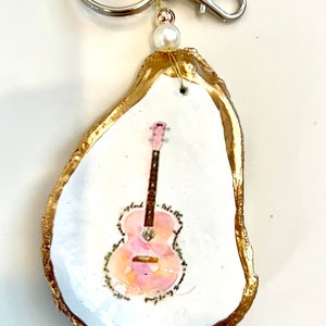 May include: A gold-rimmed oyster shell keychain with a painted pink and brown guitar on a white background.