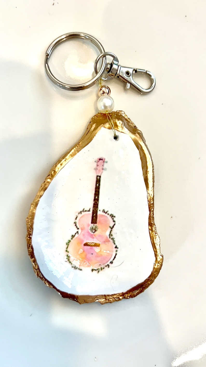 May include: A gold-rimmed oyster shell keychain with a painted pink and brown guitar on a white background.