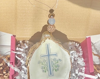Op de afbeelding: Een witte oesterschelp met een gouden rand, met een blauw kruis en witte bloemen. De schelp hangt aan een touwtje met een lichtblauwe en gouden kraal.