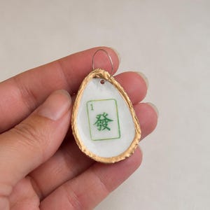 May include: A handmade pendant featuring a white, teardrop-shaped design with a gold-colored border. The pendant has a green and white Mahjong tile design with the number 1 and a Chinese character. A silver-colored loop is attached.