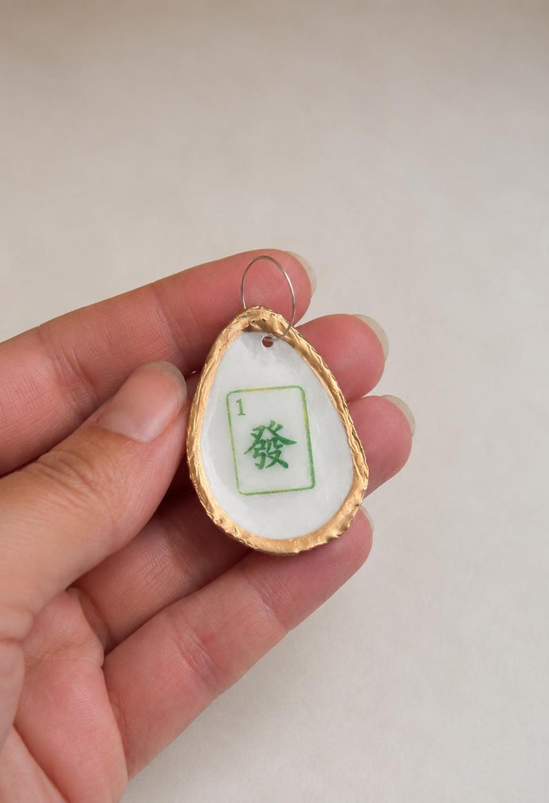 May include: A handmade pendant featuring a white, teardrop-shaped design with a gold-colored border. The pendant has a green and white Mahjong tile design with the number 1 and a Chinese character. A silver-colored loop is attached.