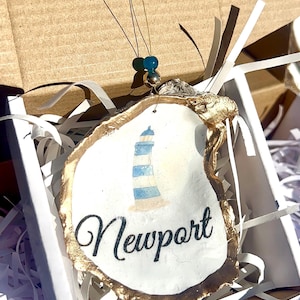 May include: An oyster shell ornament featuring a lighthouse design with blue and white stripes and the word "Newport" in black script. The shell has a gold-painted edge and is suspended by a gold string with blue and gold beads. It is presented in a white box with paper filler.