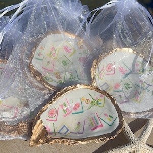 May include: Oyster shell trinket dishes with gold-painted edges, each containing a colorful design of Mahjong tiles. The dishes are presented in sheer white drawstring bags, with a starfish in the foreground.