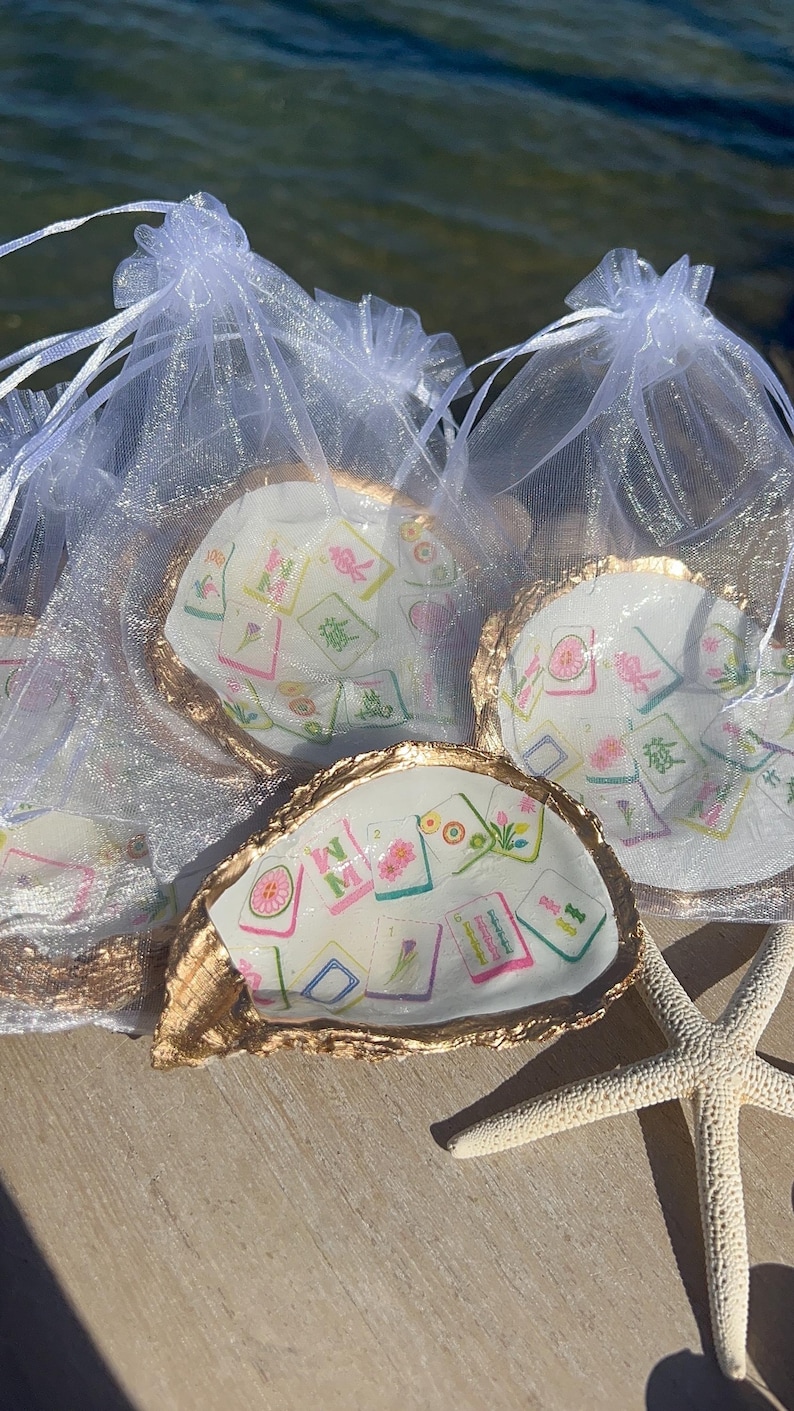 May include: Oyster shell trinket dishes with gold-painted edges, each containing a colorful design of Mahjong tiles. The dishes are presented in sheer white drawstring bags, with a starfish in the foreground.