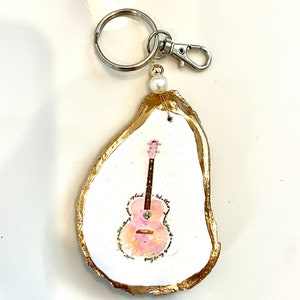 May include: A gold-rimmed oyster shell keychain with a painted pink guitar inside. The keychain has a silver ring and clasp.