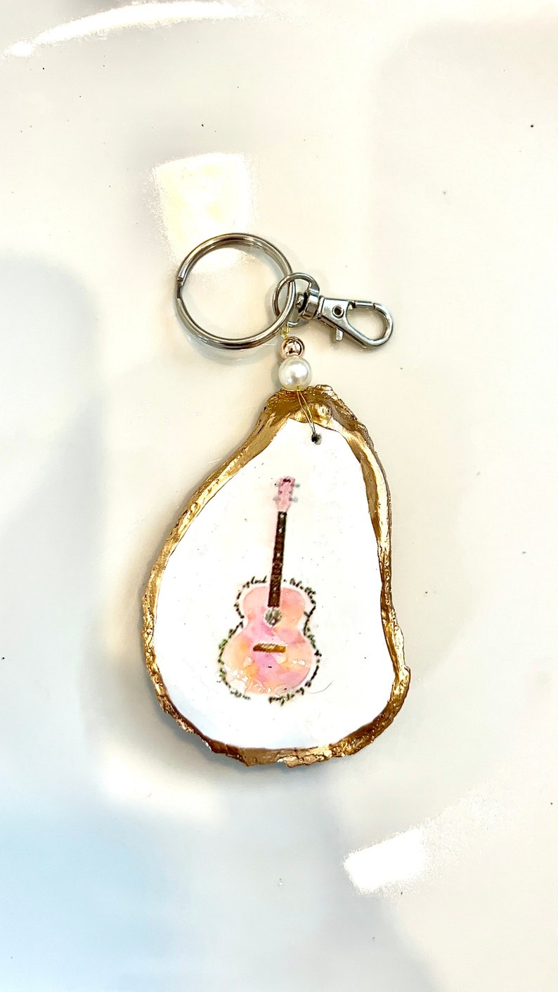 May include: A gold-rimmed oyster shell keychain with a painted pink guitar inside. The keychain has a silver ring and clasp.