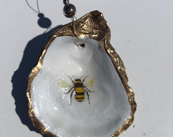 Pode incluir: Uma concha de ostra pintada de ouro com uma abelha pintada dentro. A concha está pendurada em um fio de ouro com uma conta dourada.