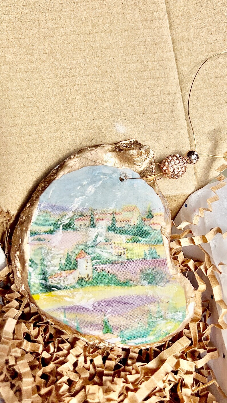 Op de afbeelding: Een handgeschilderd oester schelp ornament met een landschapssc&egrave;ne met een klein dorp, bomen en een lavendelveld. De schelp is geschilderd in tinten van blauw, groen, geel en paars, en heeft een gouden rand. Het ornament hangt aan een gouden touwtje met een kleine, glinsterende kraal.
