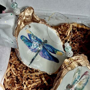May include: Two gold-painted oyster shells with a dragonfly design. The dragonflies are blue and green with white accents. The shells are hanging from a gold wire with beads.