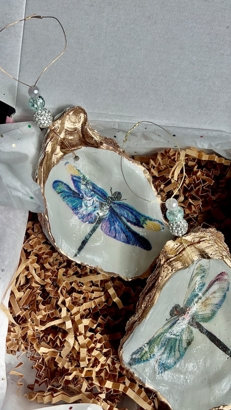 May include: Two gold-painted oyster shells with a dragonfly design. The dragonflies are blue and green with white accents. The shells are hanging from a gold wire with beads.