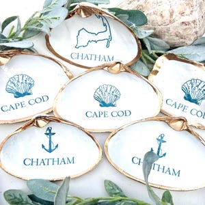 Könnte beinhalten: Sechs weiße Muschelschalen mit goldfarbenen Rändern. Jede Schale zeigt ein türkisfarbenes Design und Text: eine Muschel, ein Anker oder eine Karte von Chatham, mit den Wörtern "CAPE COD" oder "CHATHAM" darunter. Dekoratives Küstenmotiv.