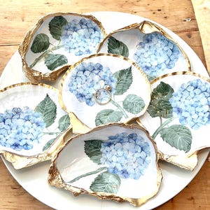 May include: A collection of five decorative oyster shells arranged on a white plate. Each shell features a hand-painted blue hydrangea flower with green leaves on a white interior, finished with a metallic gold gilded edge. A small silver ring rests in the center shell.