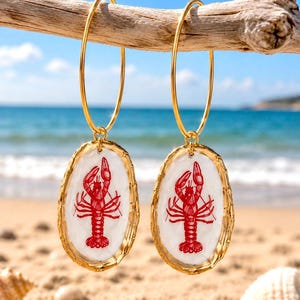 May include: Gold hoop earrings with oval pendants featuring a red lobster design on a white background. The earrings are hanging from a wooden branch, with a beach and ocean in the background.