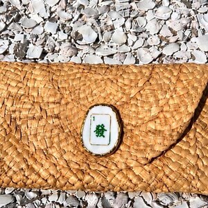 以下が含まれることがあります： フラップ開閉の織りストロークラッチ。緑色の漢字が入った白と金の装飾が施されています。バッグは長方形で、自然な薄茶色です。
