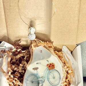 May include: A white oyster shell ornament with a gold rim, featuring a blue bicycle with a basket of flowers. The ornament is packaged in a white box with brown paper packing material.
