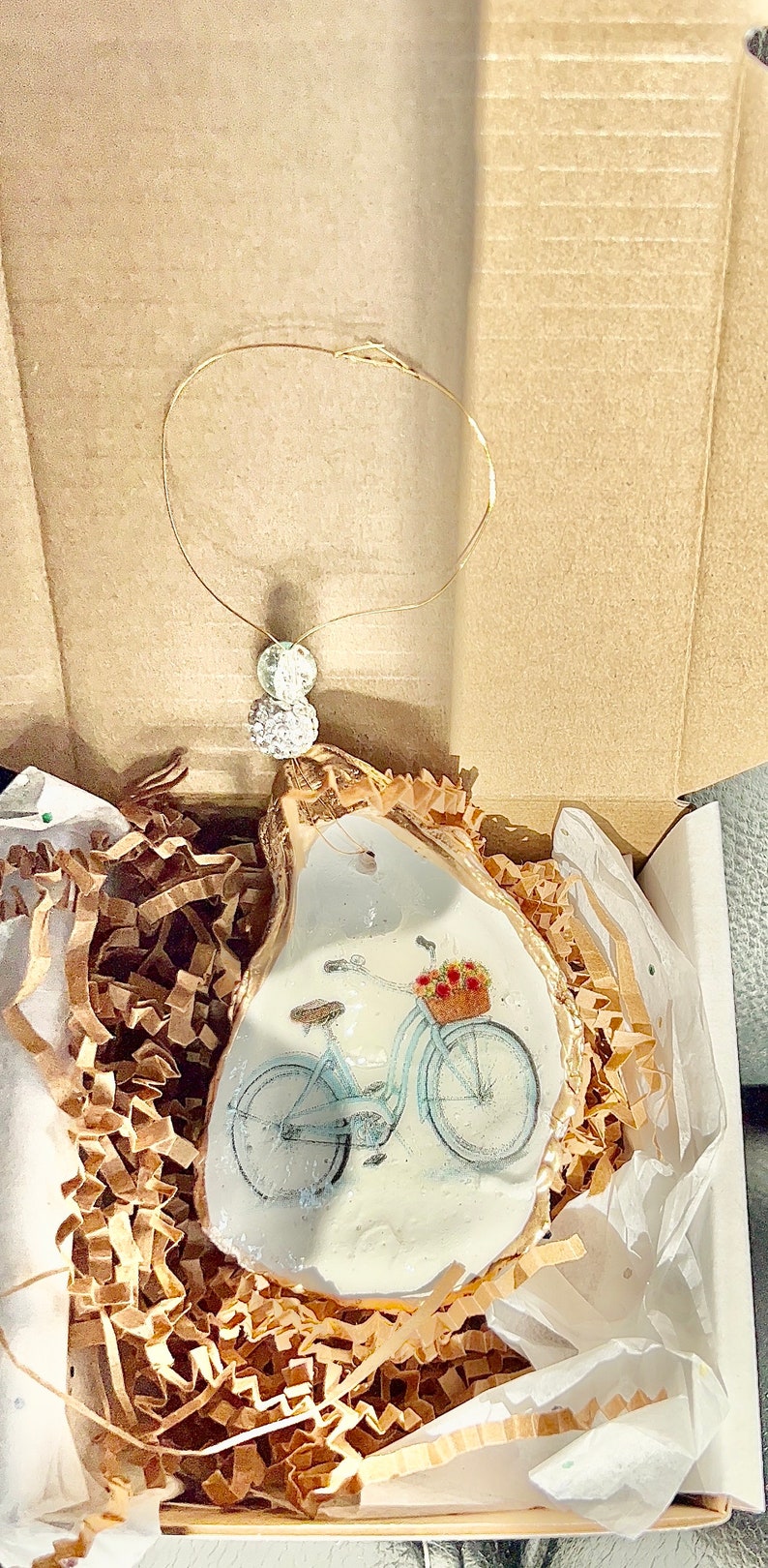 May include: A white oyster shell ornament with a gold rim, featuring a blue bicycle with a basket of flowers. The ornament is packaged in a white box with brown paper packing material.