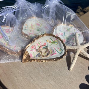 May include: Oyster shell ring dishes with gold trim, each with a unique floral design. Two gold rings with clear stones rest in one dish. Several dishes are in sheer white bags, with a starfish nearby.