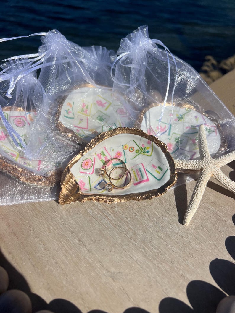 May include: Oyster shell ring dishes with gold trim, each with a unique floral design. Two gold rings with clear stones rest in one dish. Several dishes are in sheer white bags, with a starfish nearby.