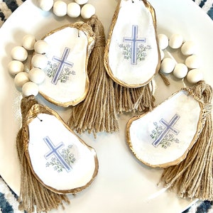 May include: Three white oyster shell ornaments with gold trim and a blue cross design. Each ornament has a brown tassel and is strung on a white bead garland.
