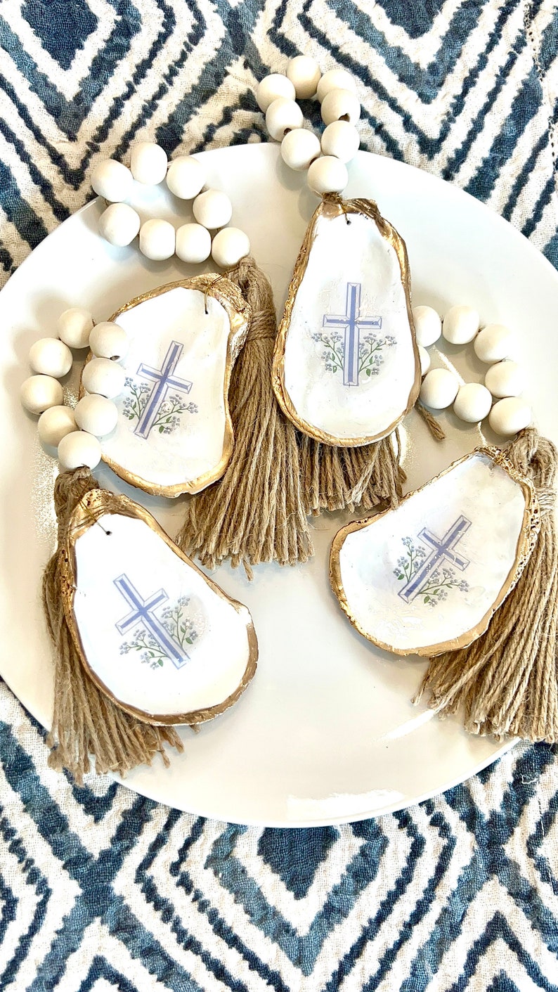 May include: Three white oyster shell ornaments with gold trim and a blue cross design. Each ornament has a brown tassel and is strung on a white bead garland.