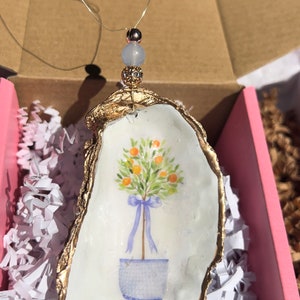 May include: A gold-rimmed oyster shell ornament with a painted image of a potted orange tree with blue ribbon. The ornament has a light blue bead and a crystal bead attached to a gold wire.