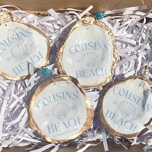May include: Four oyster shell ornaments with gold edges and the text "COUSINS BEACH NORTH CAROLINA 2023" in light blue. Each shell has a decorative bead attached for hanging. The shells are nestled in white paper filler.