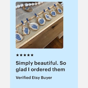 May include: A collection of handmade oyster shell pendants with blue designs and gold edges, displayed on a rectangular tray. Each pendant is unique, with a blue bead at the top. The text reads: "Simply beautiful. So glad I ordered them. Verified Etsy Buyer."