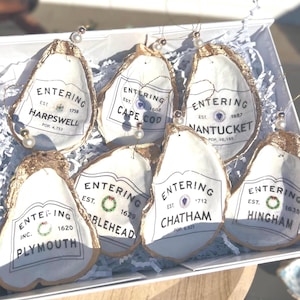 以下が含まれることがあります： 白い文字で書かれた金色のオイスターシェルオーナメント6個セット。各オーナメントには、ハープスウェル、ケープコッド、ナンタケット、プリマス、チャタム、ヒンガムなど、さまざまな町の名前が書かれています。オーナメントは小さな白いビーズで飾られています。