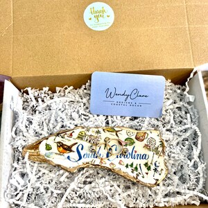 Op de afbeelding: Een witte keramische ringenschotel in de vorm van de staat South Carolina, met een gouden rand en kleurrijke illustraties van bezienswaardigheden. De schotel is gevuld met wit gescheurd papier en twee gouden ringen liggen er bovenop.