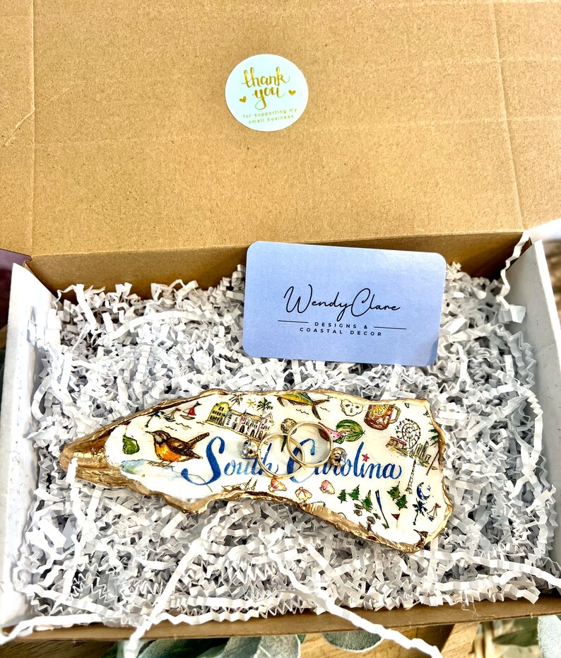 Op de afbeelding: Een witte keramische ringenschotel in de vorm van de staat South Carolina, met een gouden rand en kleurrijke illustraties van bezienswaardigheden. De schotel is gevuld met wit gescheurd papier en twee gouden ringen liggen er bovenop.