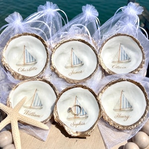 Puede incluir: Seis conchas de ostra para anillos con bordes dorados y diseños de veleros. Cada concha tiene un nombre escrito: Charlotte, James, Olivia, William, Sophia y Henry. Una estrella de mar y pequeñas bolsas blancas en el fondo.