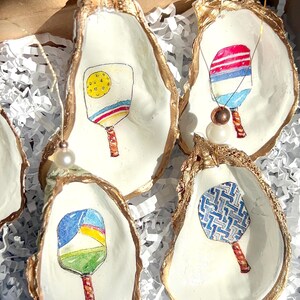 May include: Five oyster shell ornaments with gold trim, each featuring a different hand-painted design of a paddleball racket. The rackets are painted in various colors and patterns, including blue, red, yellow, green, and white.