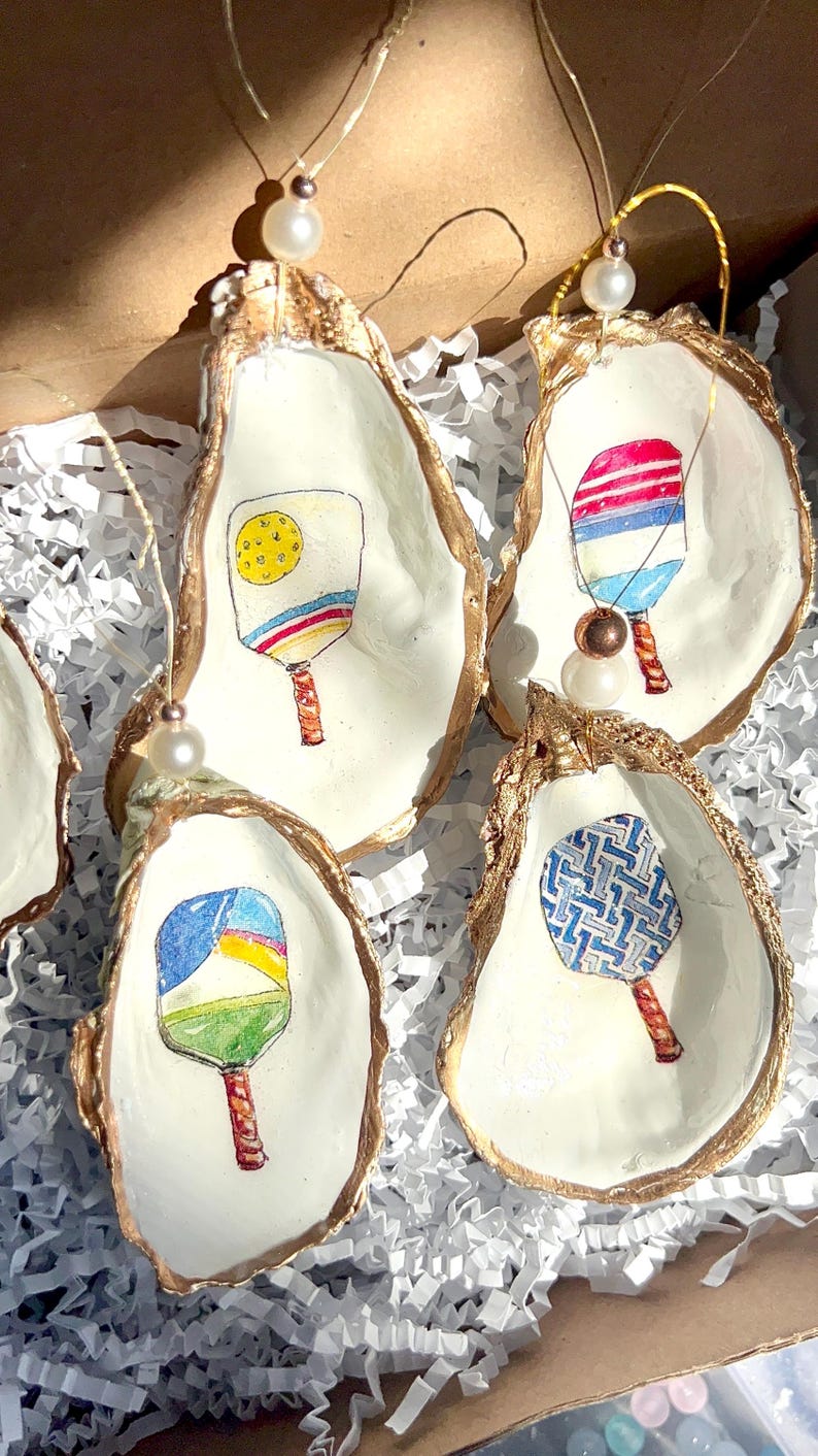 May include: Five oyster shell ornaments with gold trim, each featuring a different hand-painted design of a paddleball racket. The rackets are painted in various colors and patterns, including blue, red, yellow, green, and white.