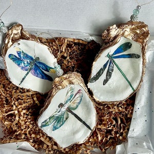 May include: Three gold-painted oyster shells with a white interior, each featuring a different dragonfly design. The dragonflies are painted in shades of blue, green, and purple.