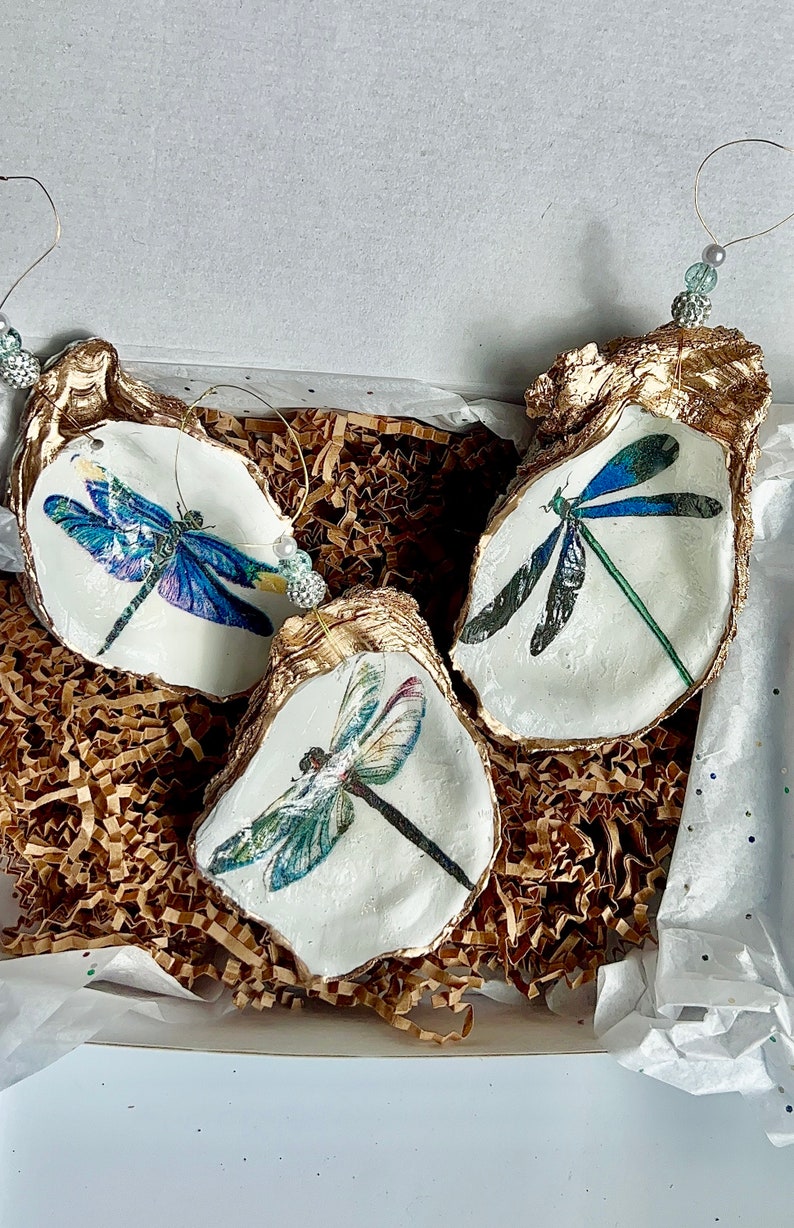 May include: Three gold-painted oyster shells with a white interior, each featuring a different dragonfly design. The dragonflies are painted in shades of blue, green, and purple.