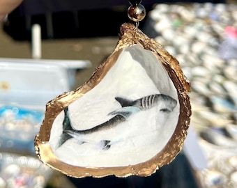 May include: An oyster shell pendant featuring a painted shark design. The shell has a gold-coloured rim and is suspended by a thin wire with a black and gold bead. The shark is depicted in shades of grey and white.