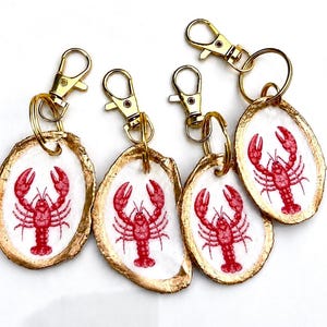 May include: Four oval-shaped keychains made from white oyster shells with gold-painted edges. Each shell features a red lobster illustration and is attached to a gold-toned metal lobster clasp and split ring for secure attachment to items.