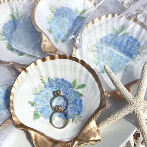 May include: Seashell-shaped ring dishes with gold trim, featuring a blue hydrangea design. Two rings rest in one dish. Several dishes are visible, some in sheer bags, with a starfish nearby.