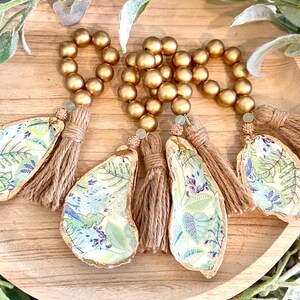 May include: Four gold beaded napkin rings with a tassel and a green and white floral design on a wooden surface.