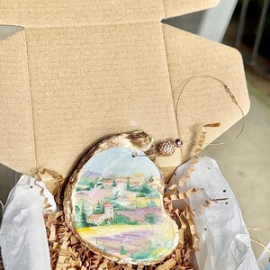 Op de afbeelding: Een handgeschilderd ornament met een landschapssc&egrave;ne met een dorp, bomen en een lavendelveld. Het ornament is gemaakt van een natuurlijk materiaal, mogelijk hout of schelp, en heeft een gouden rand. Het is verpakt in een bruine kartonnen doos met bruine papieren strooisel.
