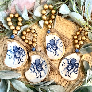 Puede incluir: Cuatro anillos de servilleta de cuentas doradas con adornos de concha de ostra pintados en azul y blanco con un diseño de pulpo. Cada adorno tiene una borla azul.